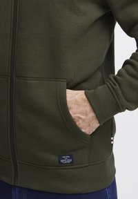 Maslinasto zelena zip-up dukserica s rebrastim manžetama, dva bočna džepa i tekstualnom oznakom na rubu. Glatka tekstura i ležeran kroj.