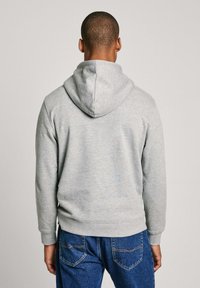 Grijze hoodie met een losse pasvorm, grote capuchon, ribgebreide manchetten en zoom. Materiaal lijkt zacht en lichtjes gestructureerd. G gecombineerd met blauwe jeans.
