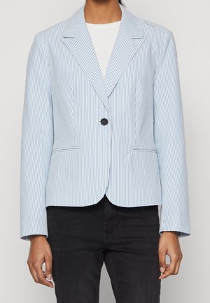 Femme portant un blazer à rayures verticales bleu clair et blanc avec un seul bouton noir, chemise blanche en dessous, et pantalon noir.