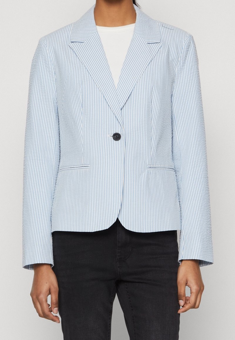 Femme portant un blazer à rayures verticales bleu clair et blanc avec un seul bouton noir, chemise blanche en dessous, et pantalon noir.
