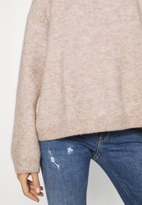 Pull en maille beige doux avec ourlet côtelé, porté avec un jean skinny bleu déchiré, affichant un style décontracté et relax de la taille jusqu'à la main.
