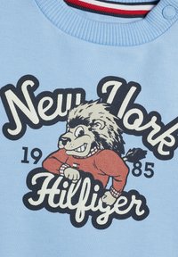 Felpa azzurra chiara con un personaggio di un leone in cartone animato che indossa un maglione rosso, con "New York" e "Hilfiger" stampati in testo nero e bianco.