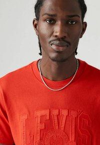 Rode katoenen T-shirt met reliëf "Levi's" logo, ronde hals, korte mouwen en een zilveren ketting. Close-up view.