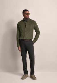 Olivgrünes Cordhemd mit Knopfleiste und Brusttasche, kombiniert mit schwarzen Hosen und hellbeigen Schuhen. Trägt eine Sonnenbrille.