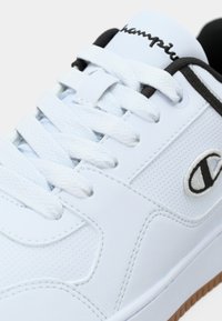 Biały sneaker z teksturowaną cholewką, płaskimi białymi sznurówkami, czarnymi akcentami i gumową podeszwą. Na boku szara logo.