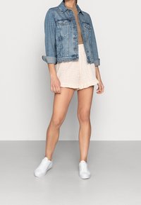 Chaqueta de mezclilla con dos bolsillos delanteros, en un lavado azul claro, llevada sobre una blusa beige y combinada con pantalones cortos de volantes a cuadros en color melocotón y blanco. Zapatillas blancas.