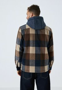 Ruuduline fliisjakke pruunis, sinises ja beežis, halli kapuutsiga; omab vaba istuvust ja nööbiga mansettide detaile.