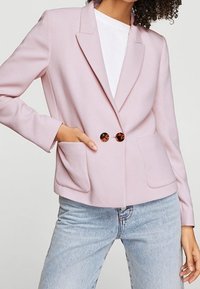 Blazer rose clair avec un design à simple boutonnage, des revers crantés, deux poches frontales et des boutons circulaires marron. Porté par-dessus une chemise blanche.