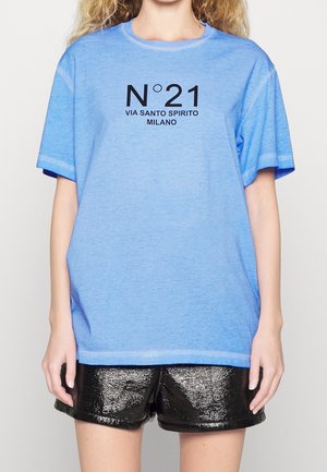 Femme portant un t-shirt bleu clair avec un texte noir "N°21 VIA SANTO SPIRITO MILANO" et un short noir brillant, debout devant un fond uni.