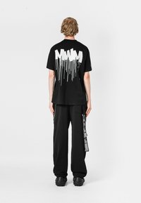 Camiseta negra con diseño de texto goteante blanco en la parte de atrás. Combinada con pantalones negros holgados que tienen bolsillos laterales y herrajes decorativos.