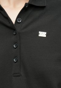 Črna polo majica iz teksturirane tkanine, s tremi črnimi gumbi in srebrno logotipno značko na levi strani.