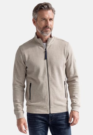 Man met grijs haar en baard, gekleed in een lichtbeige jas met ritssluiting en zwarte zakken, staand tegen een effen achtergrond.