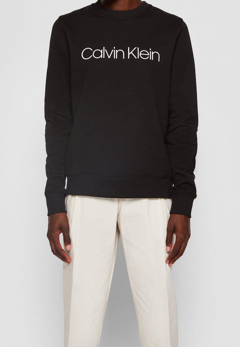 Sweat-shirt noir à manches longues avec « "Calvin Klein" » en texte blanc, porté avec un pantalon écru sur une personne debout.