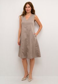 Hihaton beige mekko, jossa on V-pääntie, sivutaskut ja levenevä hame. Valmistettu sileästä kankaasta, jossa on hieno tekstuuri.