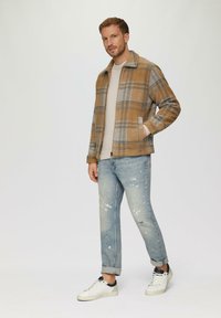 Een geblokte wollen blend jas in tan en grijs, een crème coltrui, versleten lichtblauwe jeans en witte sneakers met marineblauwe accenten.