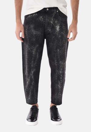 Uomo che indossa pantaloni corti maculati bianco e nero con scarpe da ginnastica nere e una maglietta bianca, in piedi contro uno sfondo semplice.