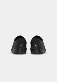 Paire de baskets basses noires vues de dos avec un petit logo rouge, blanc et bleu sur les languettes du talon sur un fond blanc uni.