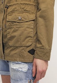 Khaki bunda s texturovaným povrchem, obsahující kapsu na patentní knoflík, nastavitelnou šňůrku a značkovou látkovou nášivku na lemu.