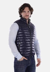 Gilet puffer blu navy con motivo trapuntato, chiusura con zip e tasche laterali, indossato sopra una camicia azzurro chiaro e abbinato a jeans scuri.