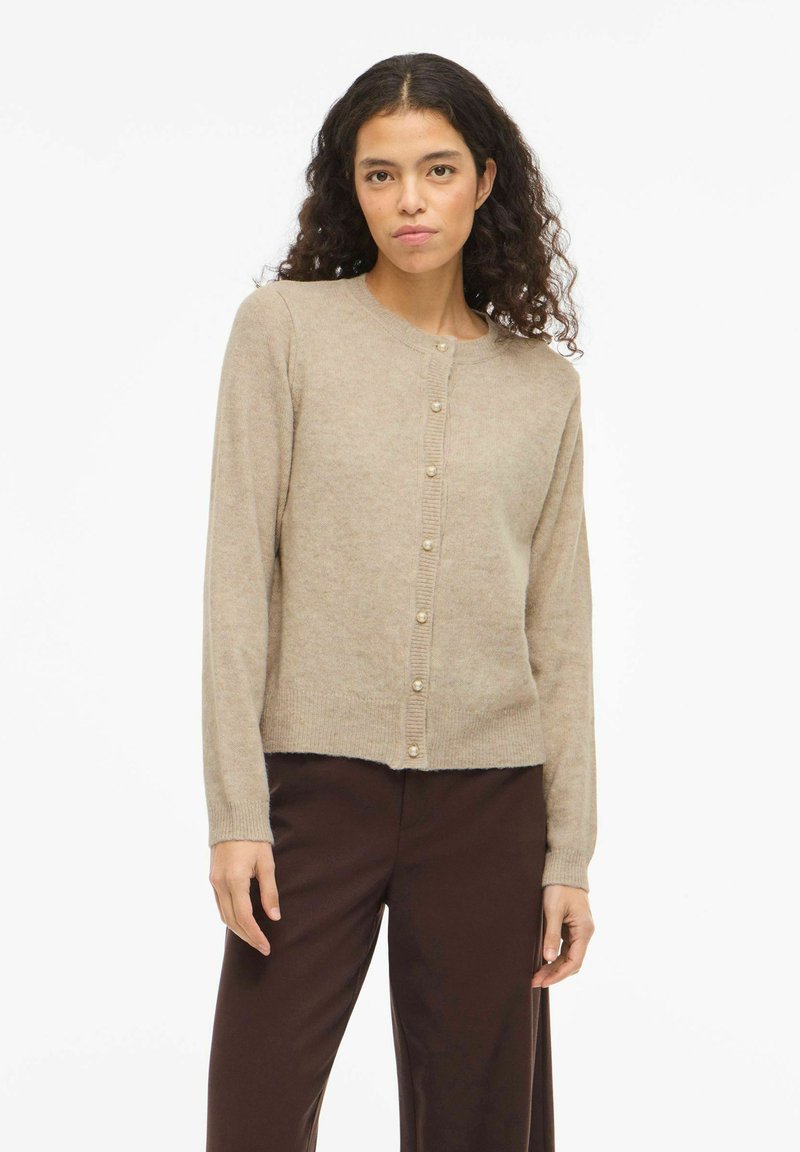 Smėlio spalvos kardiganas, pagamintas iš minkštos megztos medžiagos, su apvalia iškirpte, ilgomis rankovėmis ir perlų imitacijos sagomis priekyje. Vienodo rašto tekstūra.