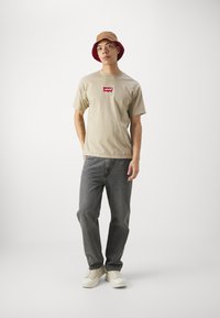 Beige bomulds t-shirt med rød Levi's-logo, grå denim jeans og cremefarvede sneakers. Bærer en lysebrun bucket hat med rød inderside.