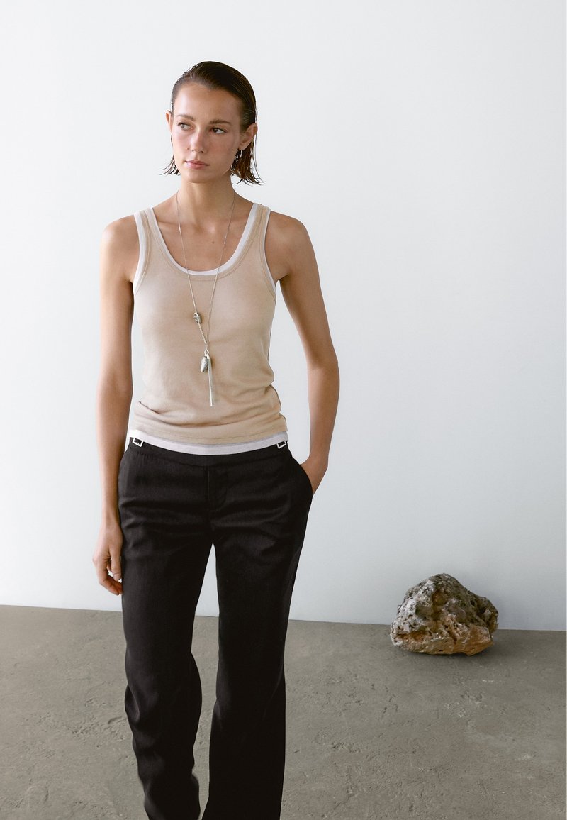 Top beige senza maniche con scollatura curva, abbinato a pantaloni neri su misura. Un semplice collier d'argento con ciondolo penzola liberamente.