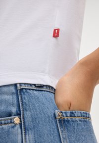 Witte katoenen T-shirt met een rood label aan de onderkant, gecombineerd met lichtblauwe denim jeans met een voorzak en gouden hardware.