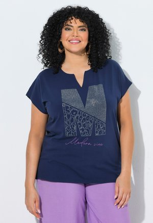 Femme souriante aux cheveux bouclés portant de grandes boucles d'oreilles créoles, un t-shirt bleu marine avec un "M" décoratif et un pantalon violet, debout contre un mur blanc.