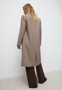 Cappotto lungo a scozzese marrone con una silhouette strutturata, caratterizzato da maniche lunghe, un unico spacco posteriore, abbinato a pantaloni a gamba larga marroni.