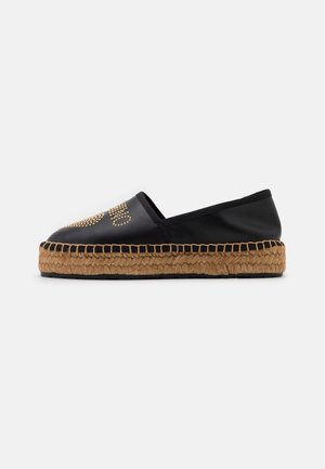 Sort slip-on espadrille med tyk flettet jutesål og guldbeslagdetaljer på overdelen.