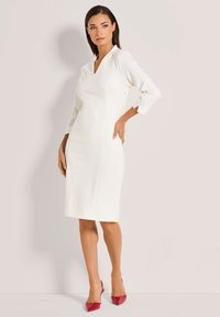 Weißes knielanges Kleid mit drei Viertelärmeln, V-Ausschnitt, glatter Textur und kontrastierenden roten spitzen Absätzen.