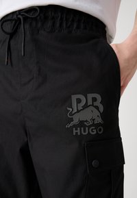 Svarte cargobukser laget av glatt stoff, med snøring i midjen, sidelomme og en stor preget logo på låret.