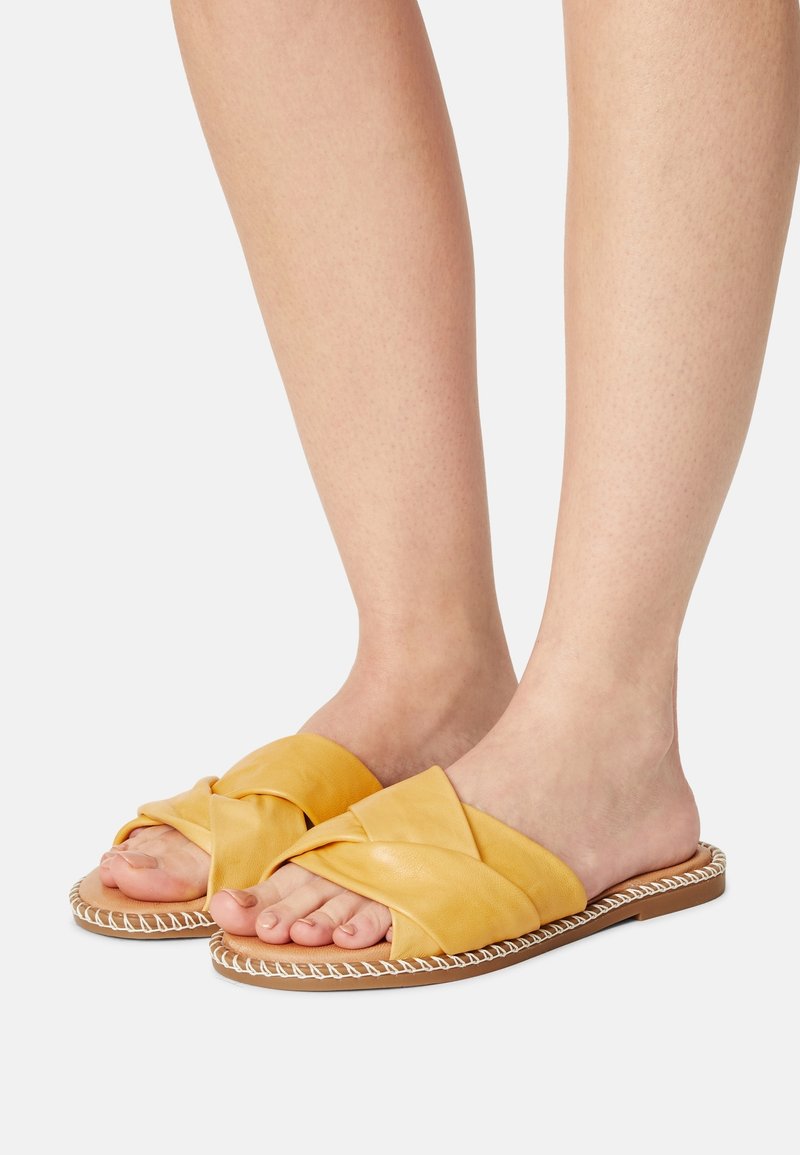 Pieds portant des sandales mule jaune moutarde avec de larges brides croisées et des semelles marron clair avec coutures blanches sur fond blanc.