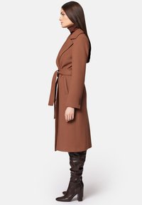 Brązowy płaszcz z wełny z paskiem, z klapami w kształcie litery V oraz dwiema kieszeniami bocznymi. W zestawie z ciemnobrązowymi wysokimi do kolan botkami. Gładka tekstura.
