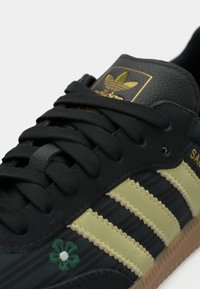 Μαύρο αθλητικό παπούτσι με πράσινες λεπτομέρειες, τρεις κίτρινες ρίγες, υφή δέρματος και λογότυπο με λουλούδι. Διαθέτει σχέδιο με κορδόνια και σόλα από καουτσούκ.