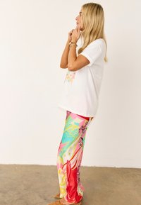 Ξανθιά γυναίκα με λευκό πουκάμισο με κοντά μανίκια και πολύχρωμο παντελόνι με σχέδια στέκεται πλευρικά σε λευκό τοίχο, ρυθμίζοντας τα κοσμήματα στο πηγούνι και τον λαιμό.