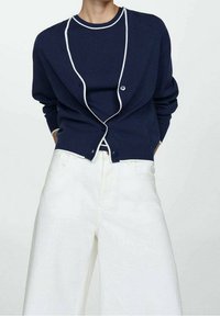 Gilet en maille bleu marine avec des bords cordés blancs, doté d'une fermeture à un bouton, porté sur un haut assorti et un pantalon large blanc.