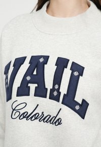 Hanorac gri cu inscripția "VAIL" brodată cu litere albastre și accente de fulgi de zăpadă albi. "Colorado" este brodat în stil cursiv dedesubt.