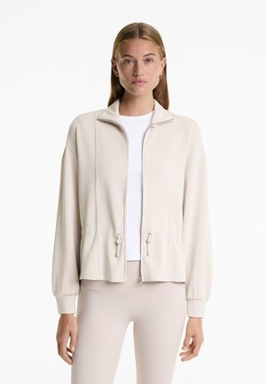 Giacca beige con zip, colletto alto, polsini elasticizzati, vita sbloccata e due toggles con coulisse, realizzata in tessuto morbido e liscio.