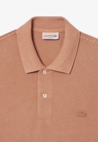 Persikofärgad polo med ribbad krage, placket med två knappar och broderad logotyp i matchande färg på vänster bröst. Strukturerad tyg.