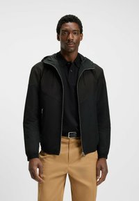Chaqueta negra con cremallera y capucha, que presenta paneles contrastantes lisos y texturizados, combinada con una polo negra y pantalones beiges.