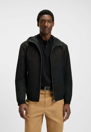 Veste noire zippée avec capuche, présentant des panneaux lisses et texturés contrastants, associée à un polo noir et à un pantalon beige.