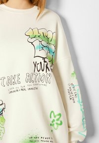 Sudadera oversize color crema con texto e ilustraciones estilo graffiti en verde y azul. Tela suave con mangas largas y escote redondeado.