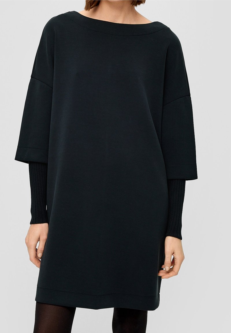 Femme portant une robe noire ample avec de larges manches jusqu'aux coudes par-dessus un haut noir ajusté à manches longues, sur un fond uni gris clair.