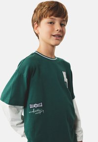 Zielona bawełniana koszulka z krótkim rękawem, z białym wykończeniem przy okrągłym dekolcie, z haftowaną literą i naszywką z napisem na rękawie.