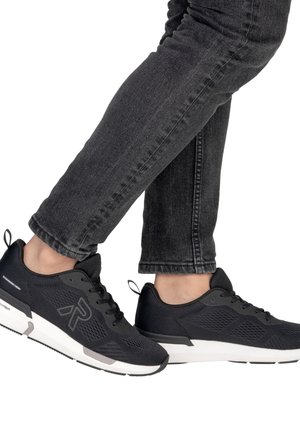 Personne portant des baskets de sport noires avec des semelles blanches et un jean gris foncé, jambes croisées aux chevilles sur un fond blanc.