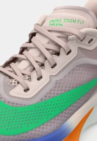 Τα παπούτσια Nike Zoom Fly διαθέτουν ελαφρύ γκρι πλέγμα από πάνω, πράσινες λεπτομέρειες και πολύχρωμο πάτο. Περιλαμβάνουν επίπεδους κορδόνια και υφή στην επιφάνειά τους.