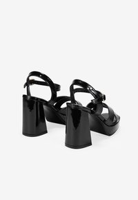 Sandalias de tacón alto de charol negro con un tacón grueso, tiras cruzadas sobre el pie y una correa ajustable en el tobillo.