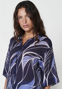 Camicia button-up di colore blu scuro con motivo a foglie in blu chiaro e bianco, con maniche corte e vestibilità rilassata, realizzata in tessuto leggero.