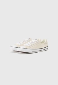 Beige suede sneakers with white laces, flat rubber soles, and black stripes along the base. Rounded toe with a smooth texture.